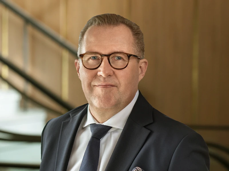 Allan Melgaard in a professional portrait, wearing glasses, a navy blazer, and a light blue dress shirt, with a confident expression