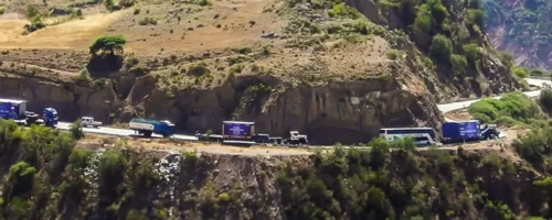 a-convoy-of-blue-semi-trucks-with-trailers-is-navigating-a-winding-mountain-road-they-are-surrounded-by-a-rugged-terrain-wi