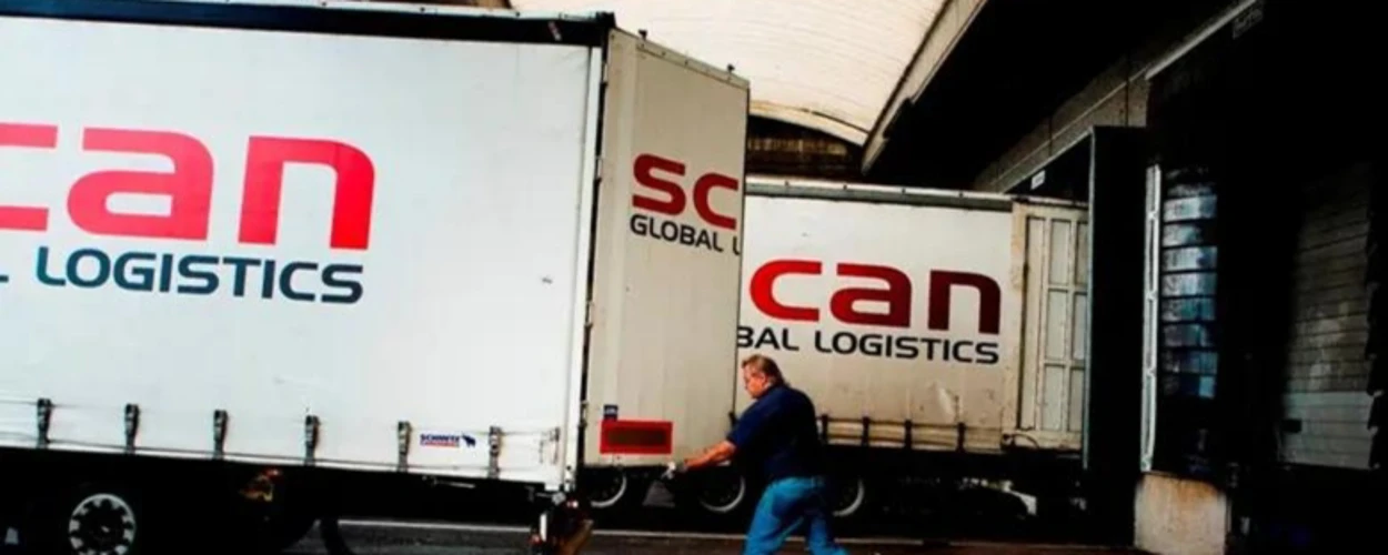 A person in blue work attire pushing a hand truck towards a truck trailer at a loading dock, with 'SCAN GLOBAL LOGISTICS' logos on adjacent trailers